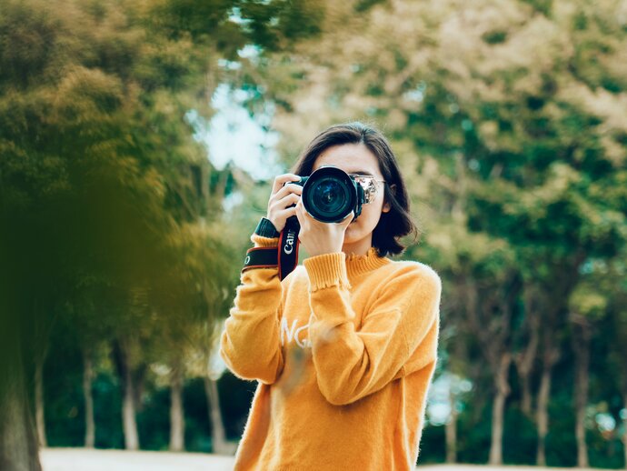 A woman in an orange jumper standing in a park with a camera held up to her face