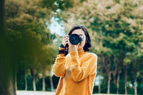 A woman in an orange jumper standing in a park with a camera held up to her face